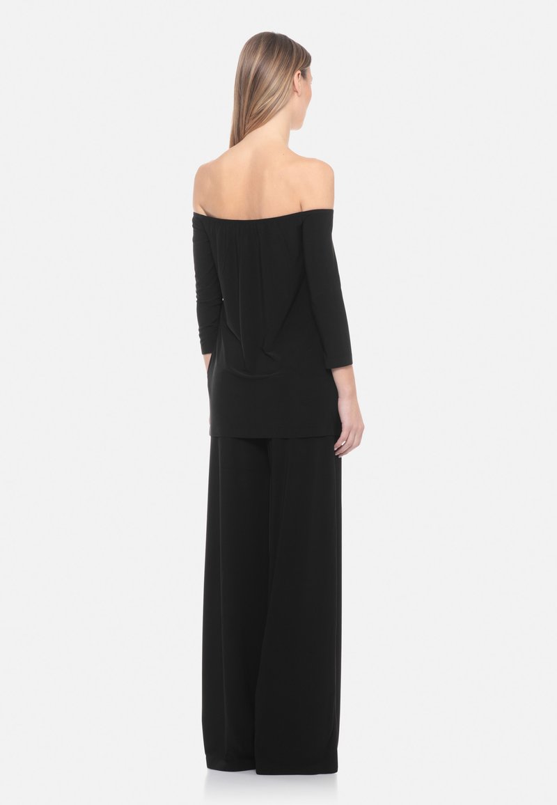 Vrouw met lang haar, gekleed in een zwarte off-shoulder top en zwarte broek met wijde pijpen, staand met de rug naar de camera tegen een effen achtergrond.