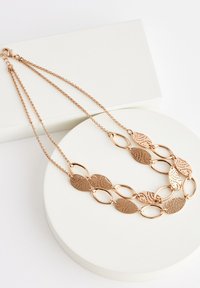 Rose gold necklace featuring interlocking leaf-shaped links with cut-out designs, suspended from dual chains, displayed on a round pedestal.