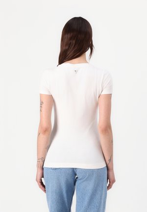 Kvinde med langt brunt hår iført en enkel hvid tætsiddende kortærmet skjorte og lyseblå jeans, stående med ryggen mod en hvid baggrund.