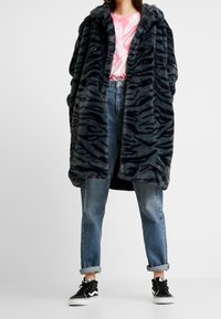 Manteau en fausse fourrure noire et grise avec un motif à rayures de tigre, doté d'un col large et d'une coupe oversize, associé à un denim bleu clair et des baskets noires.