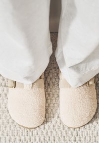 Jasnoróżowe, puchate kapcie o miękkiej fakturze. Mają beżowe, regulowane paski z metalowymi klamrami, widoczne z góry na tle teksturowanej podłogi.