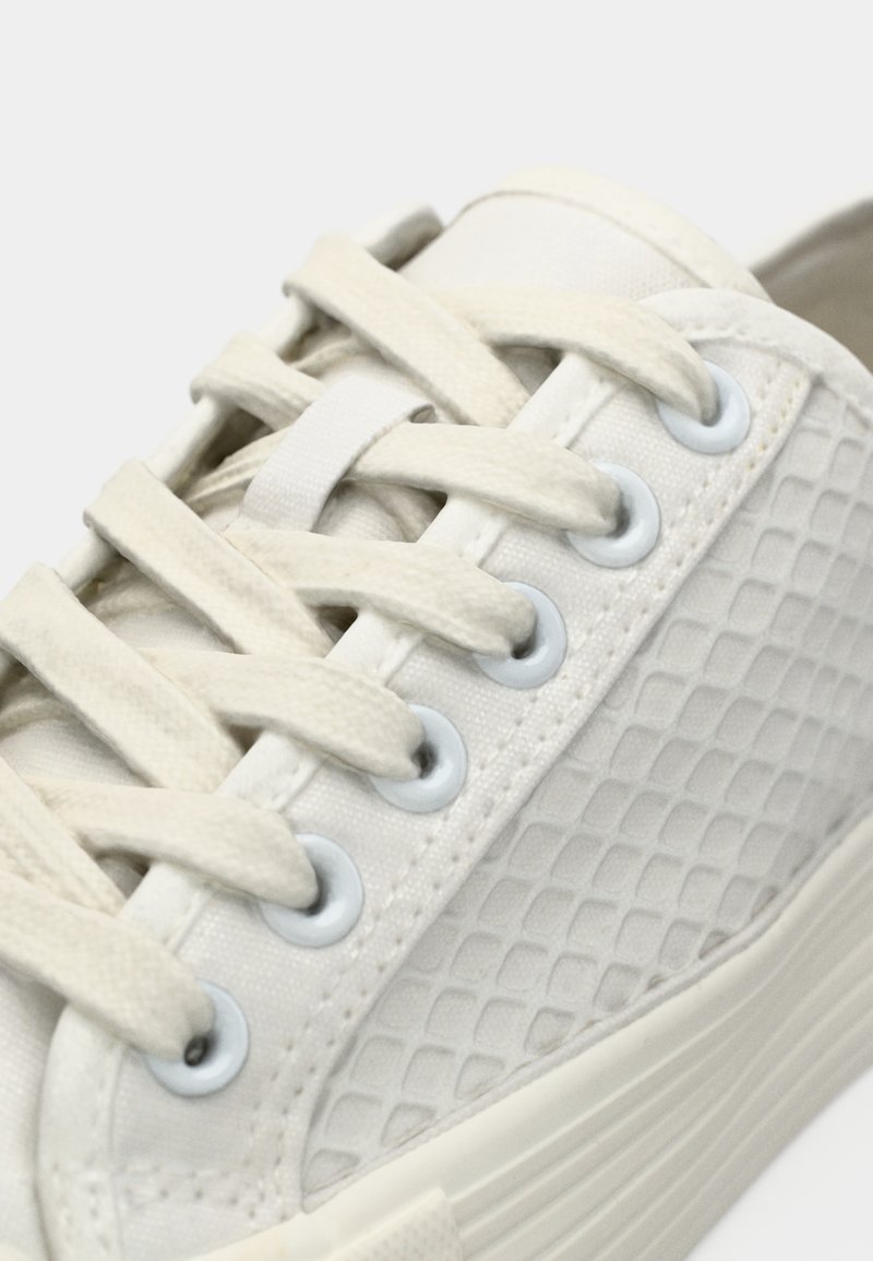 Baskets en toile blanche avec surface texturée, lacets plats, œillets bleu clair et semelle en caoutchouc, affichant un design simple et épuré.