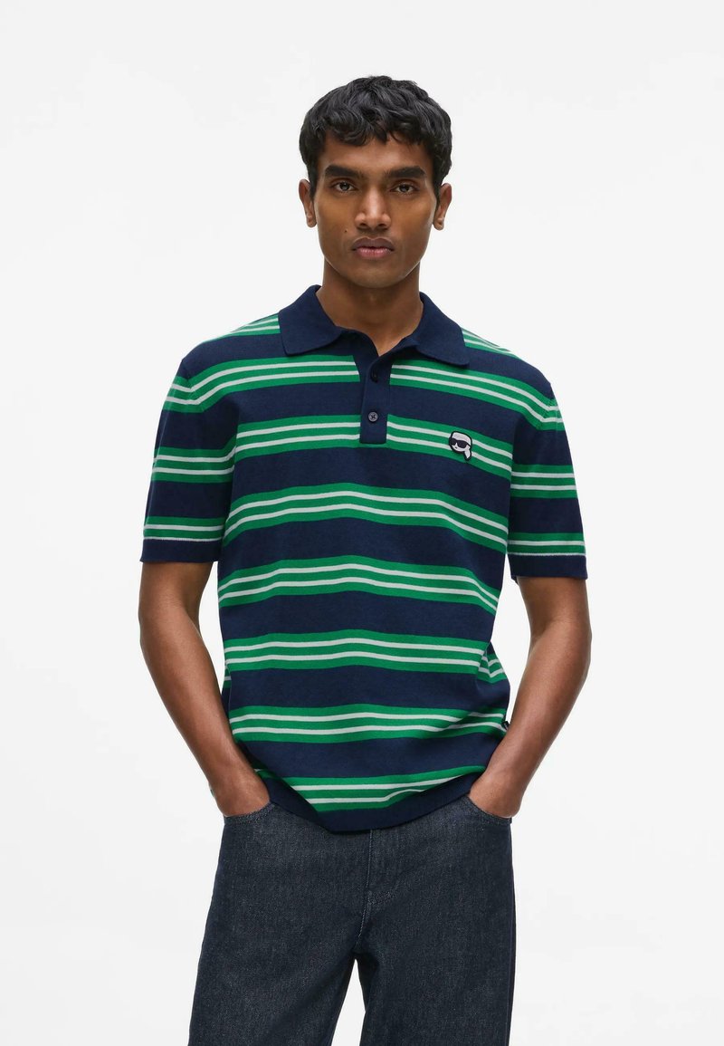 Homme portant un polo bleu marine à rayures horizontales vertes et blanches et un jean sombre, debout les mains dans les poches.