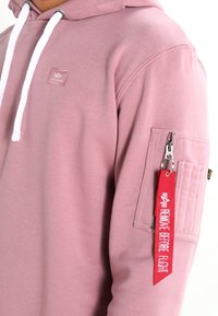 Sudadera con capucha de suave tela rosa, con cordones blancos, un bolsillo lateral con cremallera y una etiqueta roja "Retirar antes de volar".
