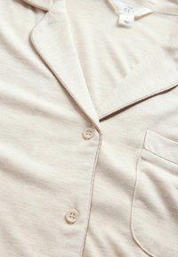Light beige pajama top made of soft fabric. Features a notched collar, front pocket, and a four-button closure. Subtle texture with light stitching.