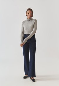 Maglione a collo alto grigio chiaro, aderente al corpo con maniche lunghe. Jeans blu scuro a gamba larga, lunghi fino alla caviglia. Scarpe basse nere.