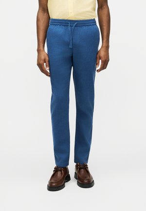 Uomo che indossa pantaloni con coulisse blu, scarpe di pelle marroni e una camicia giallo chiaro, in piedi davanti a uno sfondo bianco.