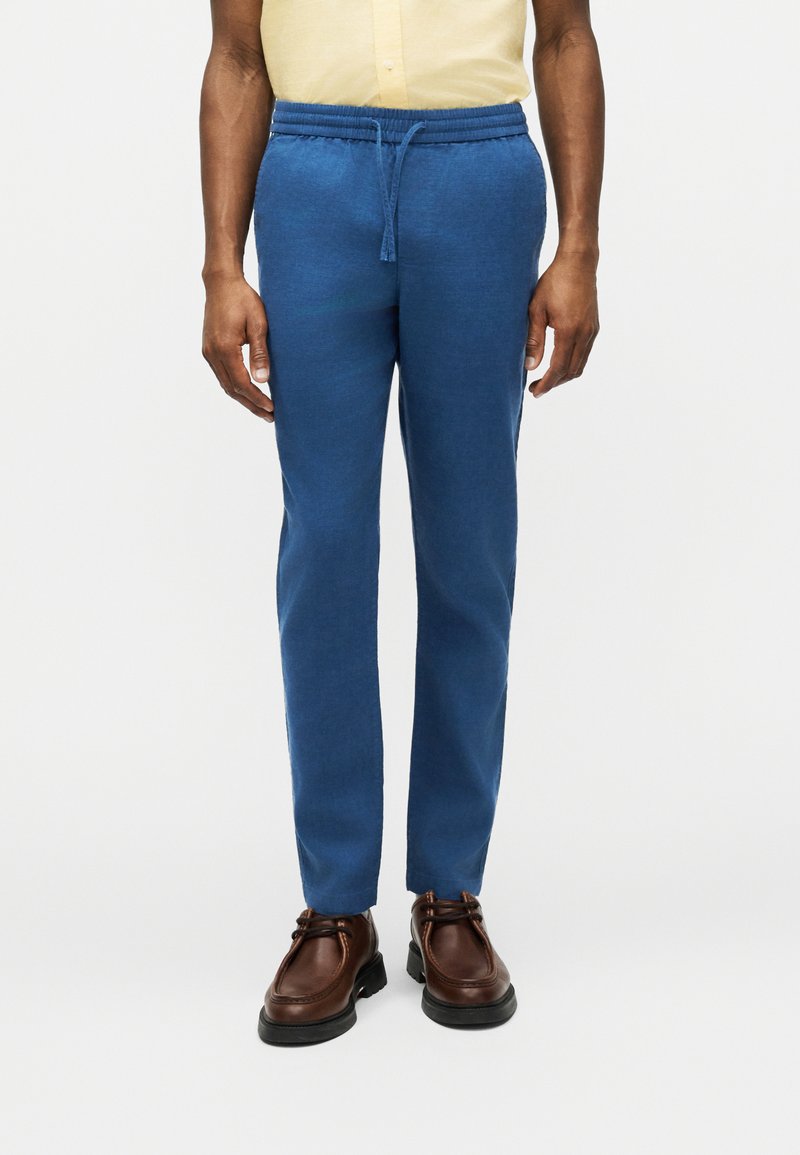 Άνδρας που φοράει μπλε παντελόνι με κορδόνι, καφέ δερμάτινα παπούτσια και ανοιχτό κίτρινο πουκάμισο, στέκεται μπροστά σε λευκό φόντο.