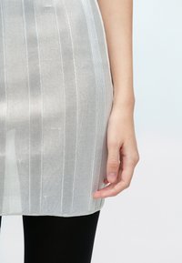 Priehľadné, pruhované šaty zo svetlosivého materiálu s lemom zakončeným nad čiernymi legínami. Detail ruky spočívajúcej na odeve.