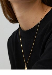 Collana d'oro con una catena fine che si posa intorno al collo, visibile sopra una maglietta nera con un collo alto. La pelle liscia è visibile sotto.
