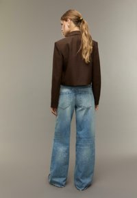Bruin cropped jasje en lichtblauwe wijde jeans. Beschadigde denimtextuur met zichtbare stiksels en achterzakken. Model staat met de rug naar de camera.