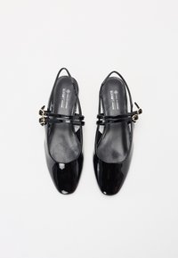 Scarpe slingback in vernice nera con punta arrotondata, due cinturini incrociati, dettagli in metallo dorato e una finitura liscia e lucida.