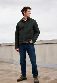 Dunkelgrüne Jacke mit braunem Kragen, blauen Jeans und braunen Schuhen. Die Jacke hat einen Reißverschluss und einen Druckknopfverschluss sowie Vordertaschen.