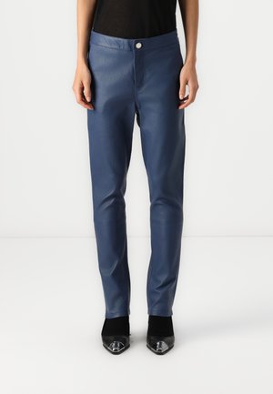 Mujer que lleva pantalones de cuero azul de corte ajustado y zapatos negros de punta afilada, de pie frente a un fondo blanco liso.
