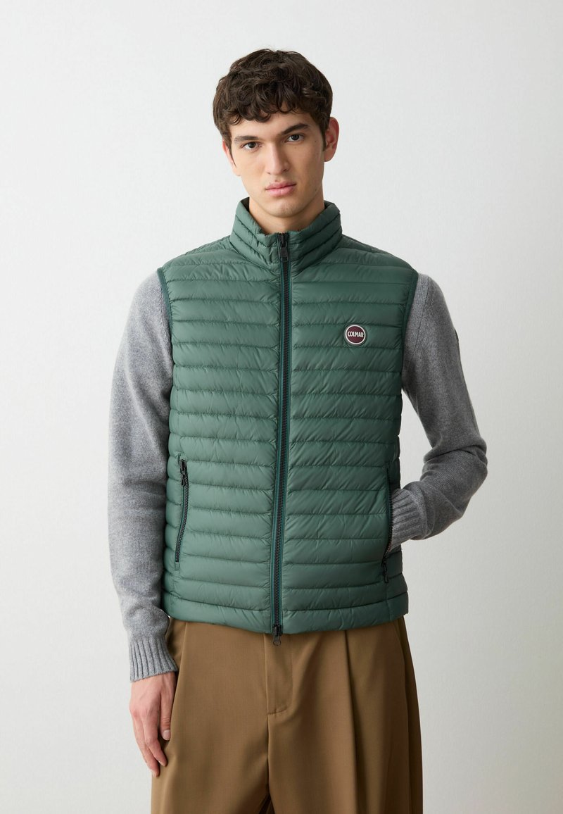 Gilet verde imbottito con colletto alto, design trapuntato, zip frontali e una patch con logo. Indossato sopra una maglietta grigia a maniche lunghe con polsini a costine.