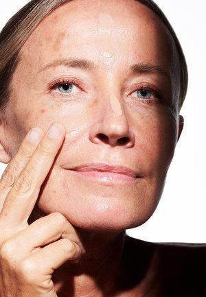 Nahaufnahme eines Gesichts mit heller Haut, das eine Creme aufträgt. Die Haut ist glatt mit wenigen Sommersprossen, und das Haar ist hell und zurückgezogen.