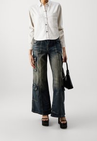 Camisa blanca corta con botones en la parte delantera, combinada con unos vaqueros anchos y desgastados de color azul que tienen bolsillos y un bolso negro. Zapatos de plataforma negros.
