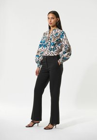Bluse mit Blumendruck in Blau- und Rosatönen, kombiniert mit schwarzer, taillierter Hose und schwarzen Riemchenschuhen, vor einem weißen Hintergrund.