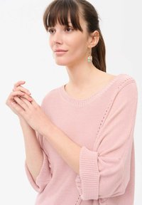 Lichtroze gebreide trui met een losse pasvorm, bootneckline en opgerolde mouwen. Het model draagt kleurrijke oorbellen en houdt haar handen samen.
