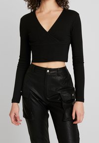 Schwarzes, geripptes langärmeliges Wickel-Crop-Top mit tiefem V-Ausschnitt, kombiniert mit hochtaillierten schwarzen Cargo-Hosen aus Kunstleder mit seitlichen Taschen.