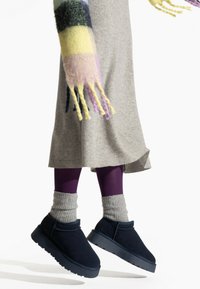 Ciemnoniebieskie zamszowe botki za kostkę z gumowymi podeszwami, połączone z szarymi wełnianymi skarpetami i fioletowymi legginsami, na jasnym tle.