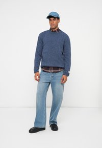 Maglione blu lavorato a maglia, camicia a quadri sotto, jeans in denim blu chiaro e scarpe nere slip-on. Un cappellino da baseball completa l'outfit.