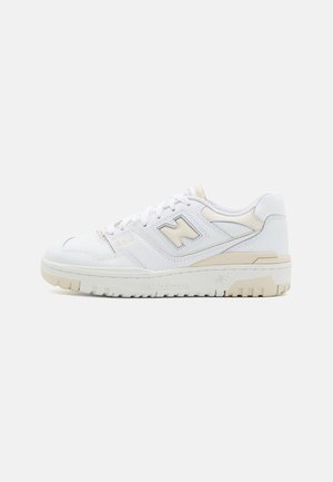 Baskets basses blanches New Balance avec accents beige, fermeture à lacets, panneau latéral perforé et semelle en caoutchouc texturée.