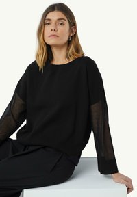 Schwarzer Pullover mit rundem Ausschnitt, einfarbigem Körper und transparenten langen Ärmeln. Die Textur ist glatt und der Schnitt ist lässig. Kombiniert mit dunklen Hosen.