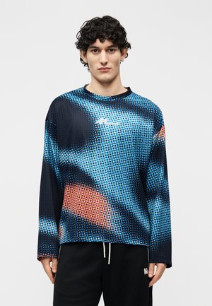 Jeune homme aux cheveux bouclés foncés portant une chemise à manches longues avec un motif abstrait de points bleu, noir et orange, et un pantalon noir avec des cordons blancs.