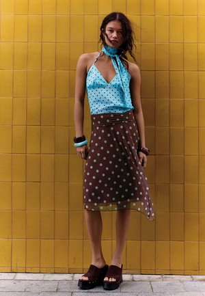 Woman stands against yellow tiled wall wearing blue polka-dot halter top, brown polka-dot skirt, platform sandals, and chunky bracelets.