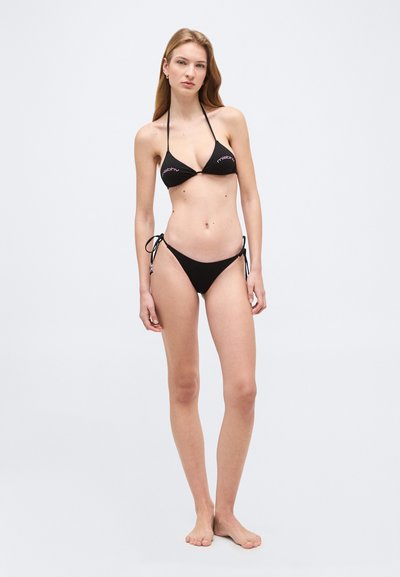 Jeune femme debout, pieds nus, portant un bikini noir en deux pièces avec un haut dos-nu et un bas à nouer sur les côtés, sur un fond uni.