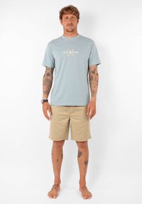 T-shirt bleu clair avec logo floral "OXBOW", associé à un short beige. Le tissu semble doux, de coupe décontractée. Modèle debout pieds nus.