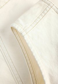 Κοντινή λήψη υφάσματος εκρού με διπλή ραφή σε μπεζ χρώμα κατά μήκος διπλωμένης άκρης, υπογραμμίζοντας την υφή και τις λεπτομέρειες της ραφής.