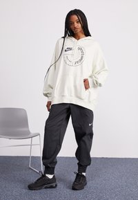 Világosszürke, oversized kapucnis pulóver fekete Nike logóval és kör alakú grafikával, fekete szűkített jogger nadrággal és fekete sportcipővel párosítva.