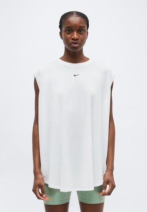 Žena na sebe má voľný biely tielkový top značky Nike a svetlozelené šortky, stojaca pred čistým bielym pozadím.