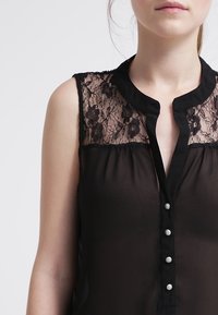 Blouse noire sans manches avec empiècement en dentelle florale, col et cinq boutons décoratifs sur le devant. Tissu transparent à la texture douce.