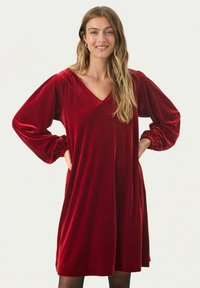 Červené sametové šaty s výstřihem do V, dlouhými nafouklými rukávy a elastickými manžetami. Splývavý design, délka ke kolenům, jemná textura a jednobarevné provedení.