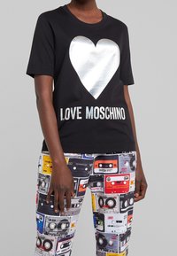 Black cotton t-shirt featuring a large silver heart graphic and "LOVE MOSCHINO" text in silver, paired with patterned multicolored cassette tape leggings.