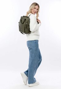 Alyvuogių žalia kuprinė su keliais užtrauktukais, sklandi nailono medžiaga, struktūrizuota forma ir dvigubomis viršutinėmis rankenomis. Modelis dėvi šviesų megztinį ir džinsus.
