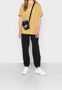 Tannfärgad bomullströja med vit "AIR"-logotyp, svarta sweatpants med elastiska nedre kanter och vita sneakers. Liten svart axelremsväska med logotyp.