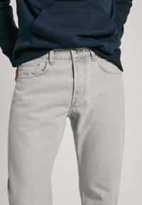 Gaiši pelēki denima džinsi ar klasisku taisno piegriezumu, pieciem kabatām un pogu aizdari, kombinēti ar tumši zilu kapuci.