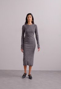 Long-sleeve, form-fitting dress made of gray textured fabric, with a below-knee length. Black, tied footwear completes the look.