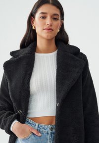 Abrigo de pelo sintético negro con un amplio cuello y detalles de botones plateados, llevado sobre un suéter blanco de punto trenzado y jeans de tiro alto.