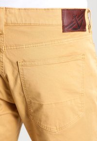 Żółte bawełniane spodnie o prostym kroju, z brązową skórzaną naszywką z motywem kotwicy i dwoma tylnymi kieszeniami.