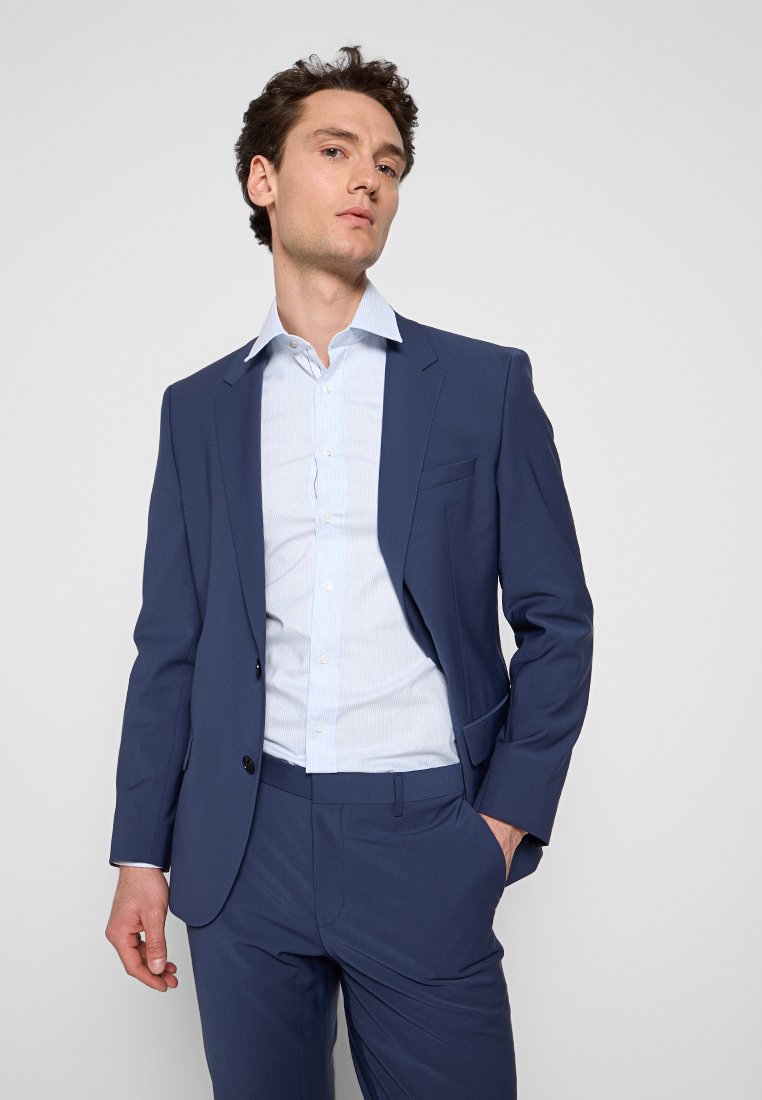 Completo blu navy con giacca aderente e pantaloni abbinati, abbinato a una camicia a righe azzurra. Tessuto liscio e design strutturato.