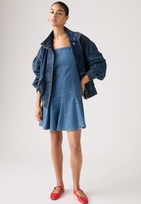 Νεαρή γυναίκα που φοράει ένα στράπλες φόρεμα από τζιν, υπερμεγέθη τζιν μπουφάν και κόκκινα παπούτσια Mary Jane στέκεται μπροστά σε ένα απλό φόντο.