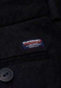 Tessuto intrecciato nero con un'etichetta "Superdry International" blu e rossa. Presenta un bottone e un motivo a tessitura testurizzata.