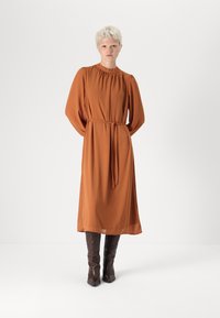 Rochie de culoare ruginie, cu mâneci lungi, decolteu cu pliseuri, talie trasă în dungi și material fluid, asortată cu bocanci de piele maro închis, până la genunchi.