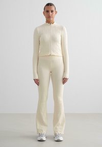 Completo a due pezzi in giallo pallido, con texture rigata; giacca aderente con zip e orlo a rouche; pantaloni a zampa abbinati; indossato con sneakers grigie.