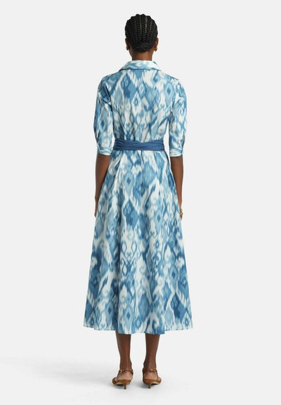 Žena s pletenými vlasmi, oblečená v modro-bielych vzorovaných midi šatách s troma štvrťovými rukávmi a hnedými sandálmi s nízkym podpätkom, zobrazená zozadu.